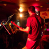 A musician plays guitar on stage under red lighting while a small audience watches in a dimly lit venue.